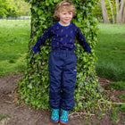 A young boy stands by a tree in EcoSplash Waterproof Fleece Lined Trousers Navy. He's wearing a long-sleeved top and wellies, looking right.